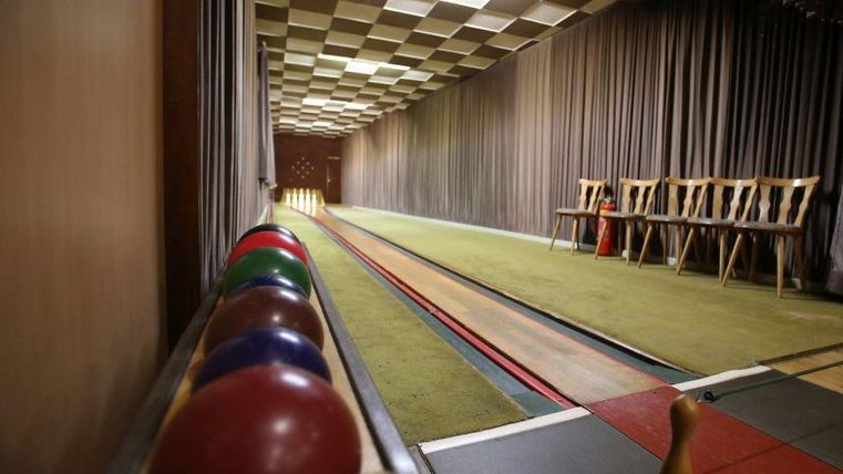 Een bowlingbaan met kleurrijke ballen en een lange, groene speelruimte. Aan de zijkant staan enkele houten stoelen.