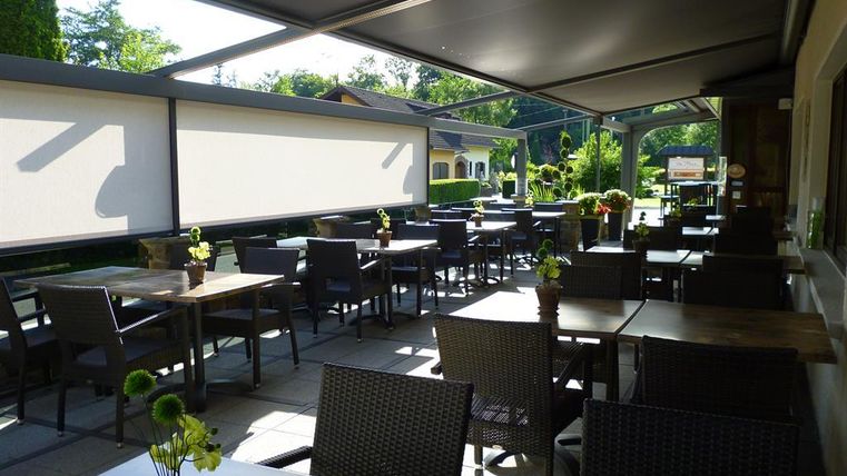 Ein gemütlicher Außenbereich eines Restaurants mit Tischen und Stühlen. Kleine Pflanzen schmücken die Tische und die Umgebung ist grün und einladend.