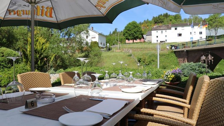 Ein einladender Tisch im Freien mit Stühlen aus Rattan, gedeckt mit Gläsern und Geschirr. Im Hintergrund sind grüne Hügel und ein charmantes Gebäude zu sehen.