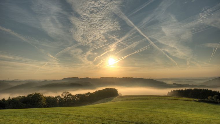 Sonnenaufgang über einer nebligen Landschaft mit grünen Feldern und bewaldeten Hügeln.