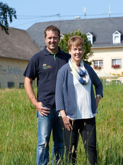 Daniela und Andreas stehen lächelnd in einem grünen Feld. Im Hintergrund ist das Hotel und ein blauer Himmel zu sehen.