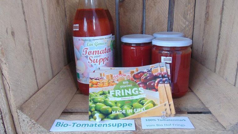 A selection of organic tomato soups in jars and bottles on a wooden shelf. Next to it is an advertisement for fresh regional products.