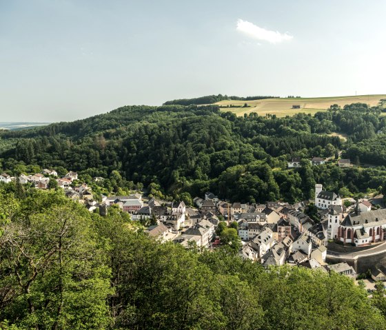 Panoramablick auf Neuerburg mit einer Kirche im Vordergrund, umgeben von dicht bewaldeten Hügeln und einem Windrad im Hintergrund., © Eifel Tourismus GmbH, D. Ketz