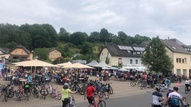 Eine belebte Platz mit vielen Menschen und Fahrrädern. Im Hintergrund sind Häuser und Bäume zu sehen, sowie Sonnenschirme.