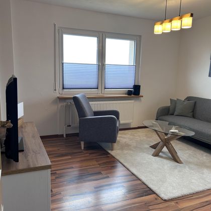 Ein modernes Wohnzimmer mit einem grauen Sofa und einem Sessel. Es gibt einen Holzfußboden und einen quadratischen Tisch aus Holz.