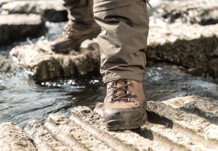 Ein Wanderstiefel steht auf einem Felsen über einem fließenden Bach. Der Stiefel ist braun und die Hose ist beige., © Eifel Tourismus GmbH, Dominik Ketz