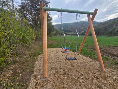 Een schommel staat op een betegelde zandbodem in een rustige park. Op de achtergrond zijn bomen en zachte heuvels te zien.