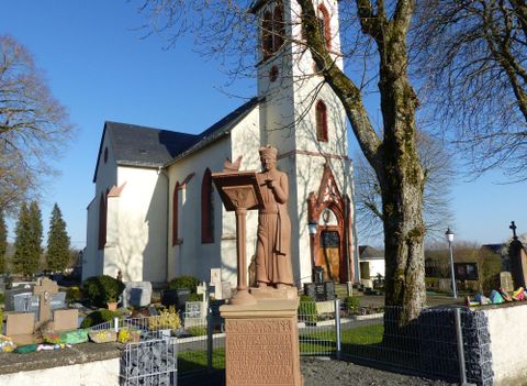 Une église avec un haut clocher et une statue au premier plan. L'arrière-plan montre un cimetière avec des arbres et un temps ensoleillé.