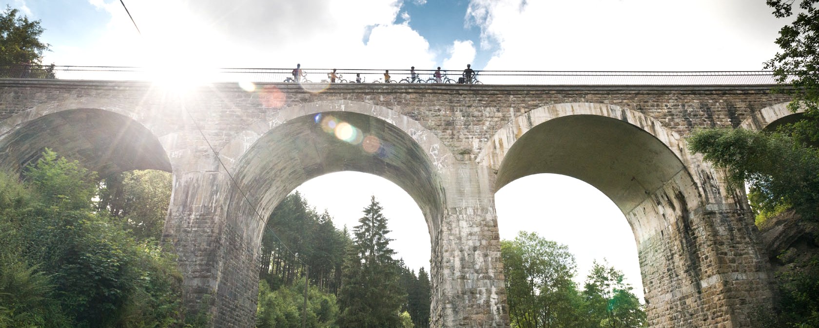 Radfahrer überqueren das Reichensteiner Viadukt der Vennbahn. Die Sonne scheint durch die Wolken, während das Viadukt über eine grüne Landschaft führt., © vennbahn.eu