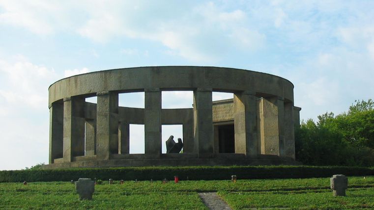 Ein markantes, rundes Denkmal mit Säulen und einer offenen Struktur. Umgeben von grünen Wiesen und Bäumen.