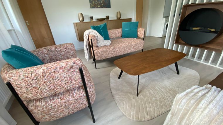 Een moderne woonkamer met twee comfortabele fauteuils, een houten tafel en een lichte kleurpalet. Op de achtergrond zijn decoratieve elementen en een tapijt te zien.