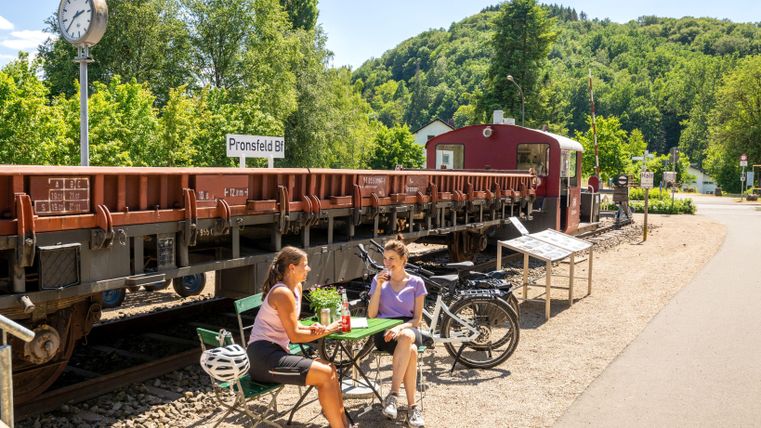 Twee personen zitten aan een tafel nabij een station. Achter hen staat een wagon en de omgeving is omgeven door groene heuvels.