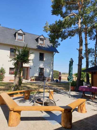 Ein gemütlicher Außenbereich mit einem Holzfeuerplatz, umgeben von Bäumen. Im Hintergrund steht ein schönes Haus mit einem blauen Himmel darüber.