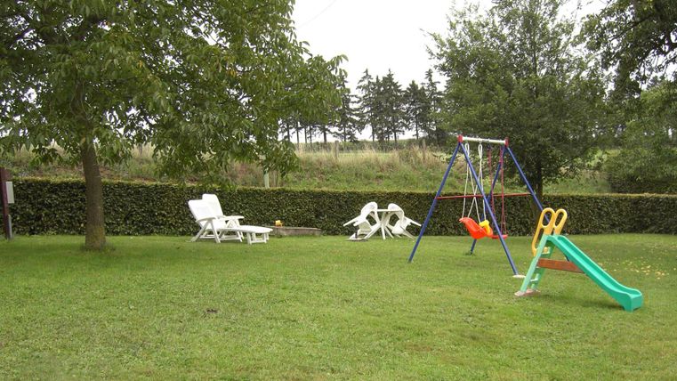 Ein grüner Garten mit einem Spielplatz, der eine Schaukel und eine Rutsche enthält. Im Hintergrund stehen Liegestühle unter einem Baum.