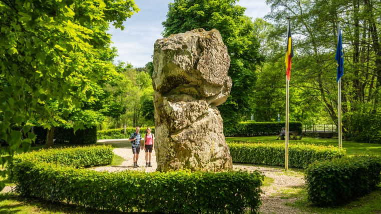Zwei Personen stehen vor einem großen Felsen im Grünen, flankiert von zwei Flaggen.