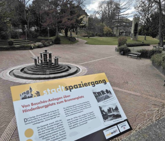 Ein Park mit einem Brunnen in der Mitte, umgeben von B&auml;umen. Ein Schild informiert &uuml;ber einen Stadtspaziergang von Bouch&eacute;s-Anlagen zum Brunnenplatz., &copy; Clara Zins-Groh&eacute;