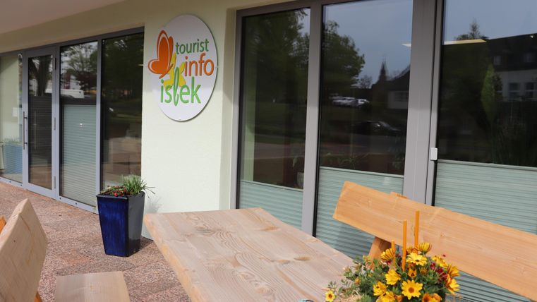 Ein modernes Informationszentrum mit großen Fenstern. Draußen steht ein Holztisch mit bunten Blumen in einem Blumentopf.