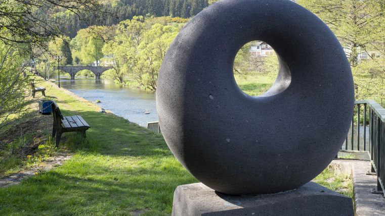 Une sculpture moderne en forme de cercle se dresse sur le rivage d'une rivière. Au loin, on aperçoit une végétation luxuriante et un pont.