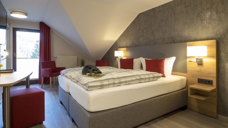 Ein gemütliches Zimmer mit einem Doppelbett, modernen Möbeln und warmen Farben. Es gibt einen Schreibtisch und einen Blick auf den Balkon.