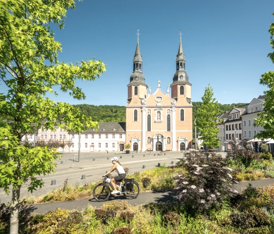 Ein Radfahrer fährt vor der Basilika in Prüm entlang, umgeben von grünen Bäumen und bunten Pflanzen, bei klarem Himmel., © Eifel Tourismus GmbH, Dominik Ketz