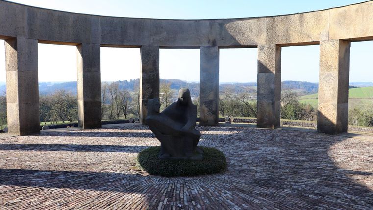Ein Denkmal in einer runden Struktur mit Säulen. Im Mittelpunkt steht eine Statue, umgeben von Natur und Ausblick auf die Landschaft.