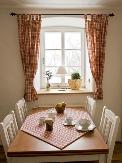 Ein gemütlicher Essbereich mit einem Holztisch, weißen Stühlen und einem karierten Tischläufer. Das Fenster lässt viel Tageslicht herein und bietet eine ruhige Atmosphäre.