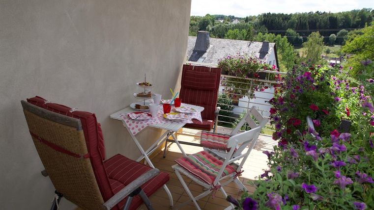 Un balcon confortable avec deux chaises et une petite table. Sur la table se trouvent une boisson et un plat à gâteau, entourés de fleurs colorées.