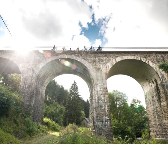 Radfahrer überqueren das Reichensteiner Viadukt der Vennbahn. Die Sonne scheint durch die Wolken, während das Viadukt über eine grüne Landschaft führt., © vennbahn.eu