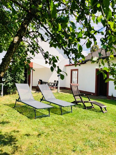 Un jardin tranquille avec deux chaises longues et une chaise. À l'arrière-plan, on peut voir une maison claire, entourée d'arbres.
