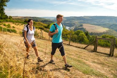 Zwei Wandernde mit Rucksäcken gehen auf einem sonnigen Pfad entlang eines Aussichtspunkts mit weitem Blick über ein bewaldetes Tal, Flusslauf und sanfte Hügel. Die Szene vermittelt Naturerlebnis und Wandern auf der Nat'Our Route.