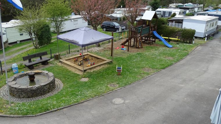 Ein Campingplatz mit Spielplatz, Sandkasten und Rutsche. In der Umgebung stehen Wohnmobile und Bäume.