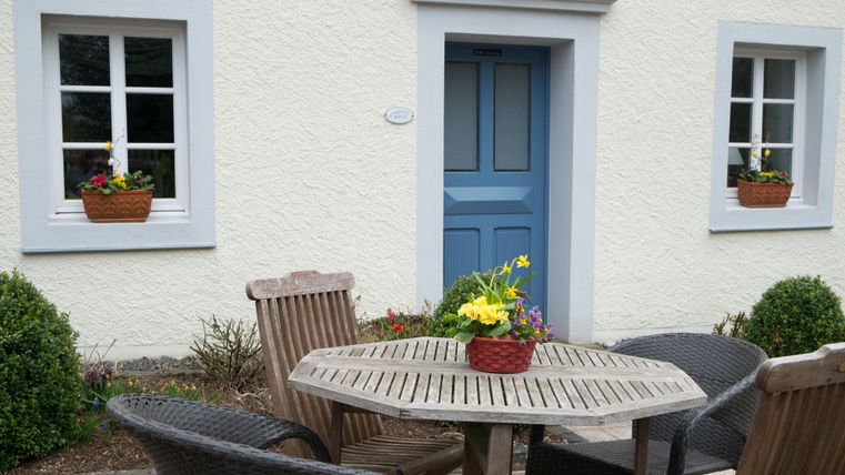 Ein gemütlicher Außenbereich mit einem runden Holztisch und Stühlen. Die Fenster sind mit Blumenkästen dekoriert und eine blau lackierte Tür zieht die Blicke auf sich.