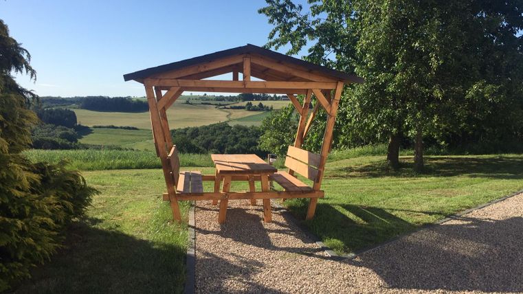 Un abri en bois avec une table et des bancs se trouve sur une prairie. En arrière-plan, on peut voir des collines douces et un ciel bleu clair.