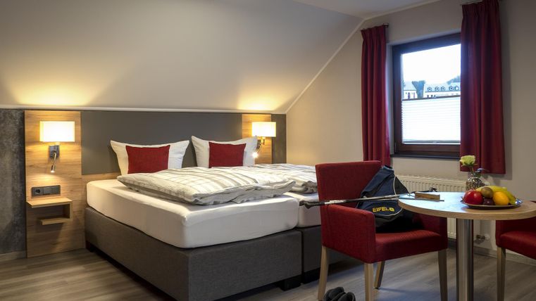 Ein gemütliches Schlafzimmer mit einem großen Bett und roten Kissen. Ein kleiner Tisch mit Stühlen und frischem Obst ergänzt die Einrichtung.