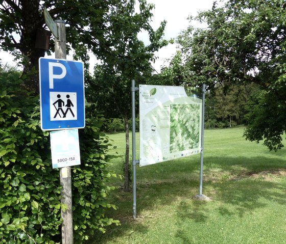 Parking lot sign for hikers next to an information board in green, wooded surroundings. Sunny day, trees and meadow in the background., © Tourist-Information Islek, Ingrid Wirtzfeld