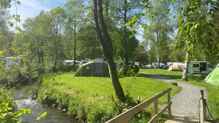Ein malerischer Campingplatz an einem ruhigen Fluss, umgeben von Bäumen und Zelten. Ein Holzsteg führt über das Gras.