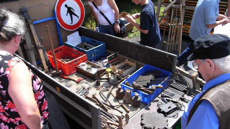 Eine Gruppe von Menschen sieht sich verschiedene Werkzeuge auf einem Tisch an. Im Hintergrund steht ein Verkehrsschild und es scheint ein sonniger Tag zu sein.