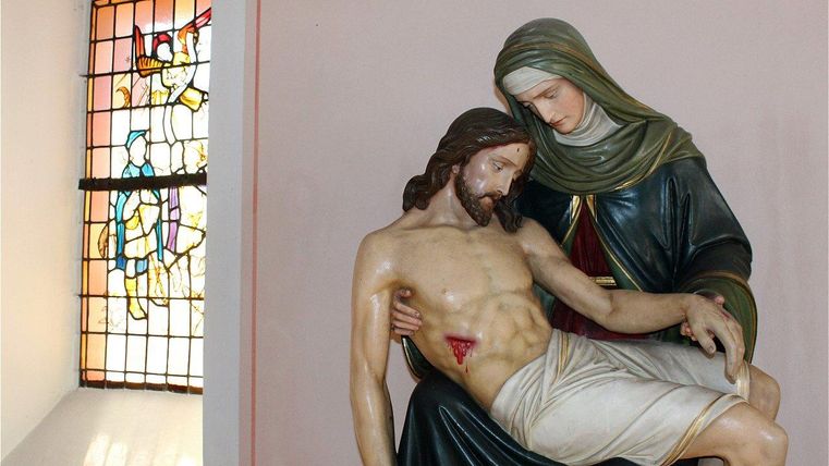 Eine Statue von Maria, die den Leichnam Jesu hält. Im Hintergrund ist ein buntes Kirchenfenster zu sehen.