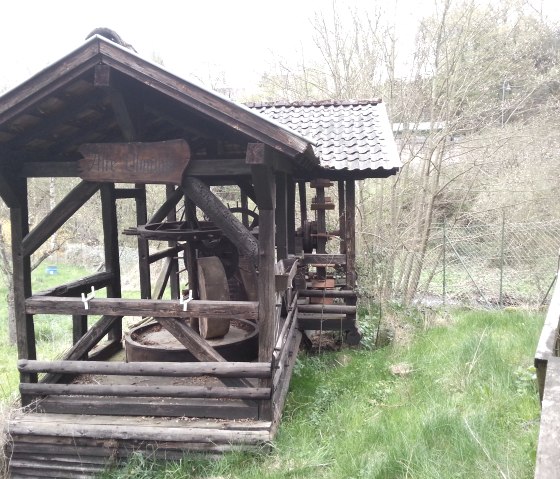 Eine alte hölzerne Ölmühle mit einem kleinen Dach steht in einer grünen, bewachsenen Umgebung., © Ti Hocheifel-Nürburgring