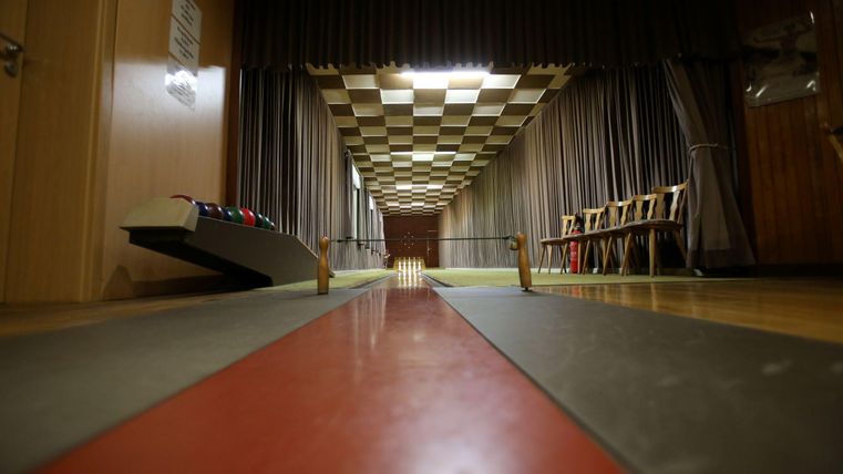 Eine Kegelbahn mit einem roten und grauen Laufbereich. Im Hintergrund sind Stühle und Vorhänge zu sehen.