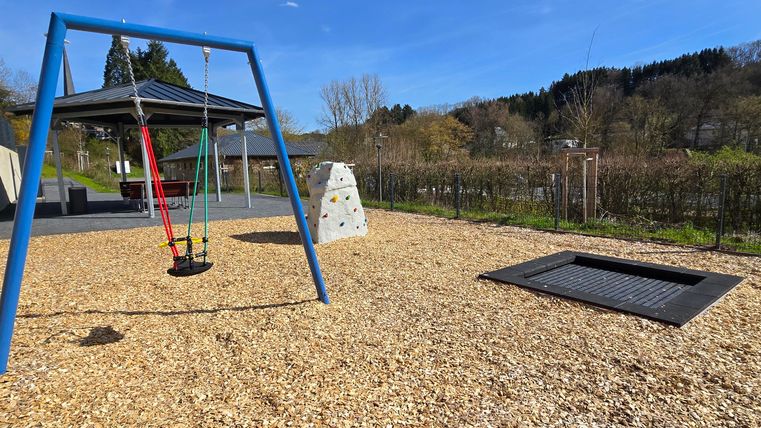 Ein schöner Spielplatz mit einer Schaubel, einer Kletterwand und einem Trampolin. Der Himmel ist klar und es gibt viel Platz zum Spielen.
