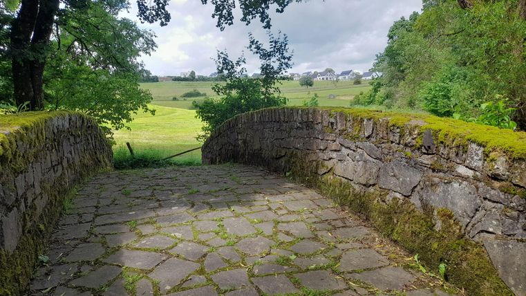 Eine steinerne Brücke führt über ein grünes Feld. Umgeben von Bäumen, zeigt der Himmel eine Mischung aus Wolken und Licht.