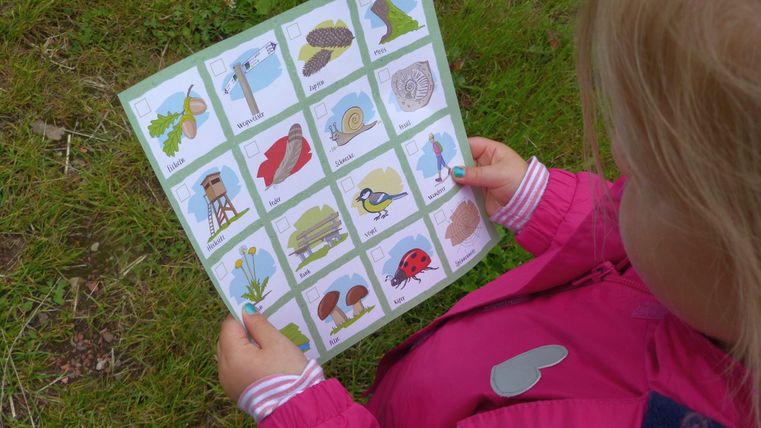 Ein kleines Kind hält eine Karte mit verschiedenen Naturmotiven in der Hand. Die Karte zeigt Tiere und Pflanzen, die in der Umgebung zu finden sind.