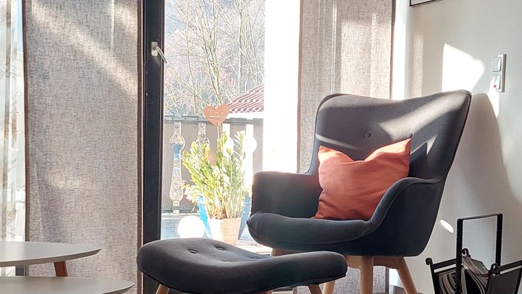 A stylish living area with a comfortable armchair and a matching footstool. Large windows let in plenty of light and provide a view of the outdoors.