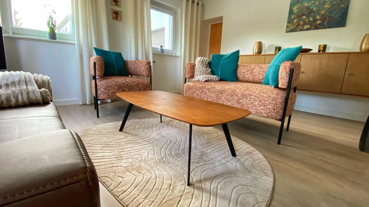 Een gezellige woonkamer met twee comfortabele fauteuils en een lage houten tafel. De muren zijn helder geschilderd, en er zijn grote ramen met gordijnen.