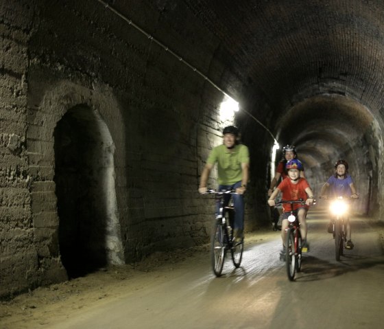 Drei Radfahrer, darunter ein Kind, fahren durch einen beleuchteten Tunnel. Der Tunnel ist Teil des Eifel-Ardennen-Radwegs, ehemals eine Bahnstrecke., © Eifel Tourismus GmbH/intention
