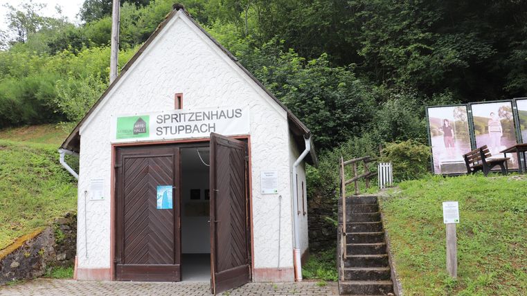 Ein kleines, weißes Gebäude mit offener Tür, das als "Hirtenhaus Stupbach" gekennzeichnet ist. Es liegt an einem bewaldeten Hang und hat Treppen, die zu einem weiteren Bereich führen.