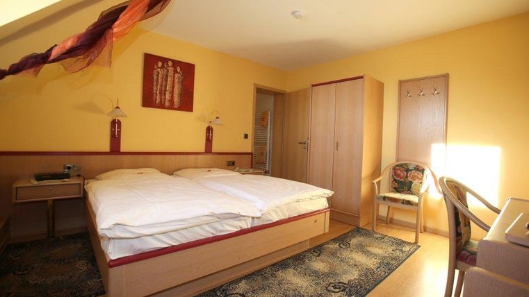 Een gezellige kamer met een tweepersoonsbed en warme gele muren. Er is een bureau, een stoel en een garderobe.