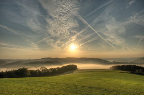 Sonnenaufgang über einer nebligen Landschaft mit grünen Feldern und bewaldeten Hügeln.