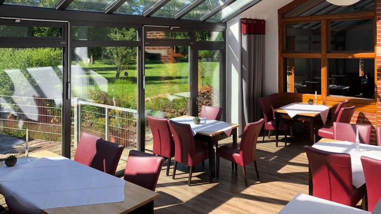 Ein helles Restaurant mit großen Fenstern und rotem Stuhl. Tische sind gedeckt und der Blick geht ins Grüne.